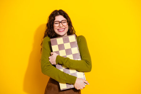 Cheerful young woman in glasses hugging vintage board game on vibrant yellow backgroundの写真素材