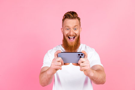Excited red-haired man with a beard using a smartphone against a vibrant pink background in casual...