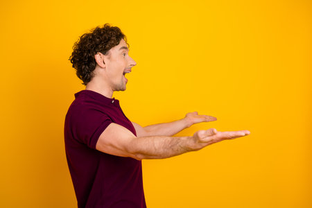 Excited young man with curly hair gesturing enthusiastically while wearing a burgundy polo shirt, standing against a yellow backgroundの写真素材