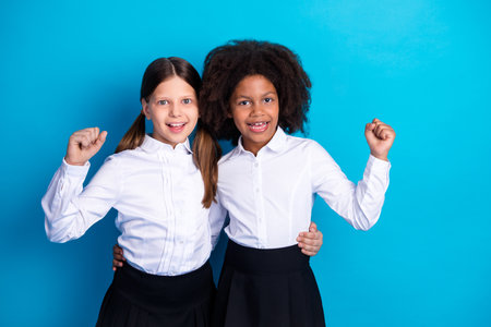 Two cheerful schoolgirls in uniforms celebrating a joyful moment while standing against a plain blue background.の写真素材