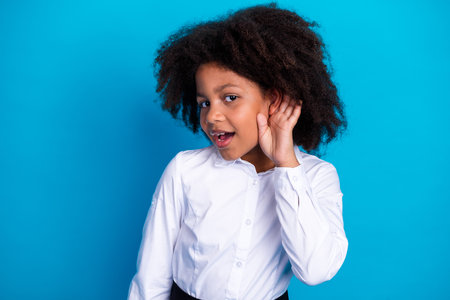 Curious young girl holding her hand to her ear, dressed in a white shirt, standing against a vibrant...