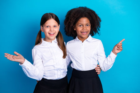 Two cheerful schoolgirls in uniforms standing against a blue background, smiling warmly while gesturing expressivelyの写真素材