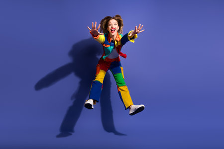 Energetic young woman in colorful outfit playfully jumps with a bright smile against a vibrant purple backdropの写真素材
