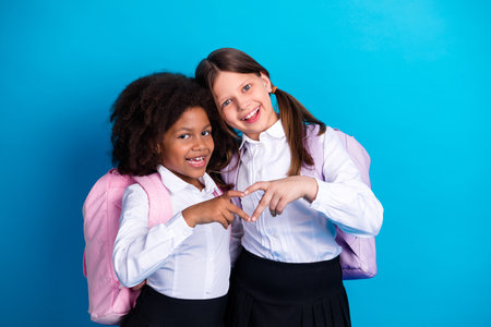 Two diverse schoolgirls in uniforms smiling with playful gestures against a bright blue background, showing joyful friendship.の写真素材