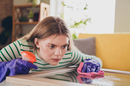 Young woman in casual attire cleaning a table while wearing rubber gloves in a cozy living room with...