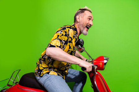 Charismatic middle aged man rides festive red scooter against vibrant green backdrop, embodying summer fun in playful printed shirtの写真素材