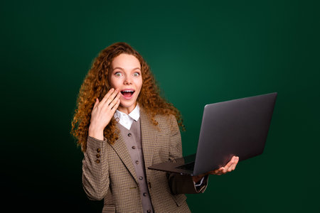 Confident young businesswoman holding laptop, expressing surprise, wearing elegant formal attire against emerald green backgroundの写真素材
