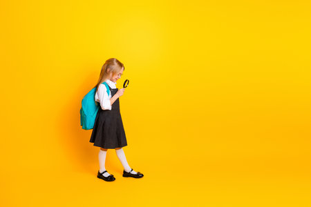 Smart schoolgirl with backpack holding magnifying glass among bright yellow background learning and...