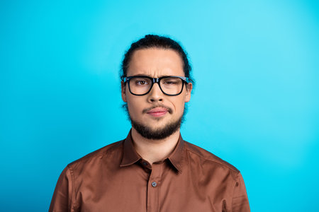 Young man with glasses displaying a skeptical look against a turquoise background, wearing a brown shirt in formal styleの写真素材