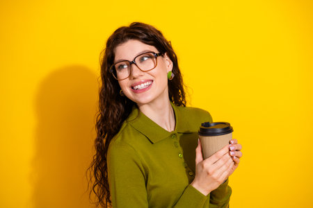 Charming woman holding a coffee cup against a vibrant yellow background, smiling joyfully and dressed in a green shirtの写真素材
