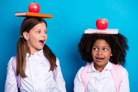 Two cheerful schoolgirls balance book with apples on their heads against a blue background, showcasing education and funの写真素材