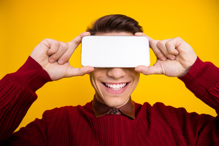 Young Man Holding Smartphone Over Eyes in Front of Bright Yellow Background