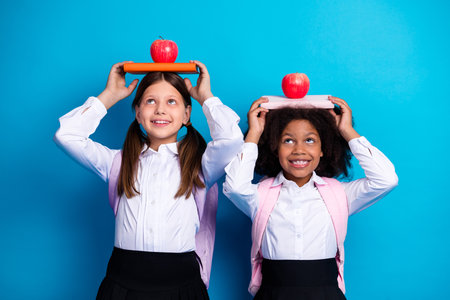 Two cheerful schoolgirls balancing book and apples on their heads, showcasing their joyful learning and studying momentsの写真素材