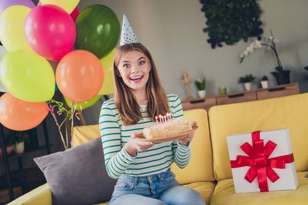 Cheerful young woman celebrating birthday at home with balloons, a cake, and a wrapped giftの写真素材