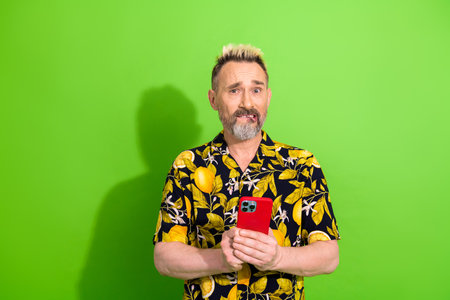 Stylish mature man with dyed hair holding a smartphone against a green background, wearing a vibrant...