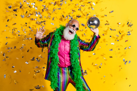 Photo of extravagant funny aged man dance disco ball confetti wear shiny costume isolated on yellow...