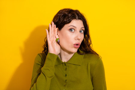 Young woman making a curious listening gesture with hand raised in front of a bright yellow...