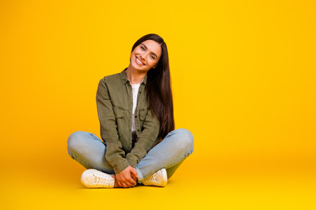 Young woman seated on yellow studio background in casual attire showcasing happiness and relaxed fashion lifestyleの写真素材