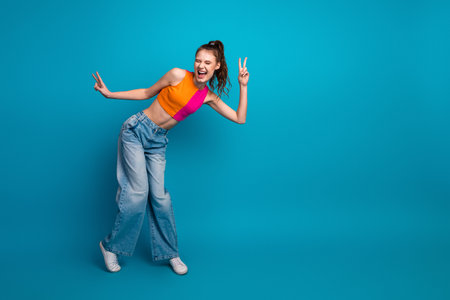 Cheerful young woman in vibrant casual outfit making peace sign against a bright blue backgroundの写真素材