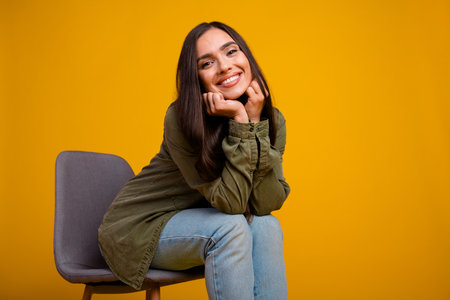 Young woman in casual attire sitting and smiling with joy against a bright yellow background, emphasizing positivityの写真素材