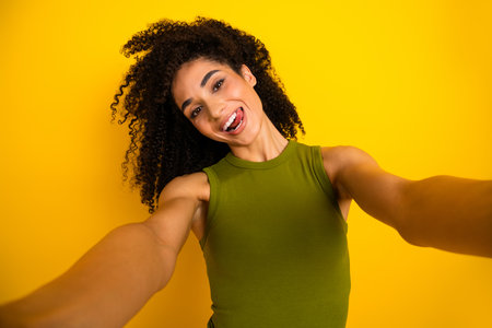Happy young girl in green top posing for selfie with curly hair on vibrant yellow background...