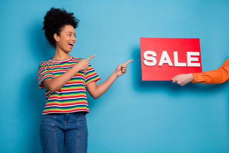 Young woman in colorful striped shirt pointing at bright sale sign, promoting discounts against...