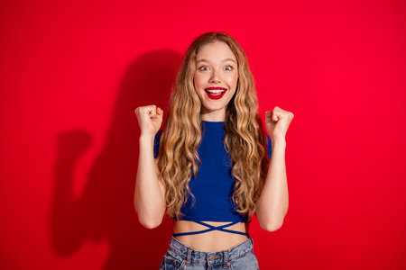 Joyful young woman with long blonde hair, wearing a blue top, enthusiastically expresses happiness against a vibrant red backgroundの写真素材