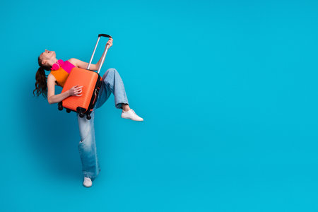 Energetic young woman in casual attire with suitcase posing against vibrant blue background exuding fun travel vibesの写真素材