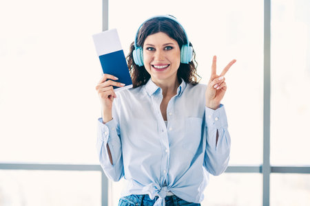 Happy young woman with passport and headphones showing a peace sign while at the airport, excited for a vacation.の写真素材