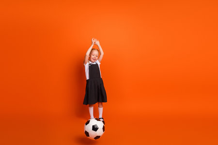 Adorable schoolgirl playing football on vibrant orange background, expressing joy and charm, dressed in a casual school uniformの写真素材