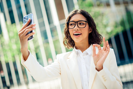 Confident young businesswoman making an engaging gesture during a video call outdoors in a modern urban settingの写真素材