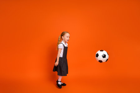Happy young schoolgirl playing with soccer ball against vibrant orange background in playful atmosphereの写真素材
