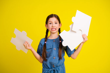 Adorable brunette girl holding large puzzle pieces on a bright yellow background, expressing confusion, in casual styleの写真素材