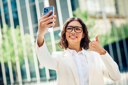 Confident Young Businesswoman Taking a Selfie Outdoors with Thumbs Up in Modern City Environmentの写真素材