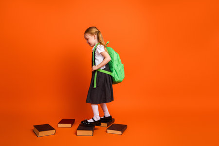 Cheerful young girl in school uniform stepping on stacked books, against vibrant orange background, promoting educationの写真素材