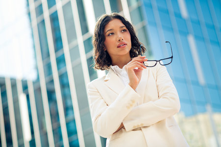 Successful businesswoman in formal attire holding glasses against a modern city backdrop, exemplifying successの写真素材