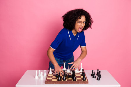 Young woman in blue polo shirt showing an expressive gesture while playing chess on a pink studio backgroundの写真素材