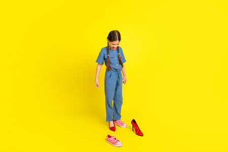 Adorable young girl with braids standing against bright yellow background wearing a blue jumpsuit and pink sneakersの写真素材