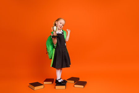 Young schoolgirl celebrates achievement in winning a trophy with enthusiasm surrounded by books on an orange backgroundの写真素材