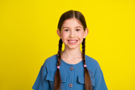 Smiling young girl wearing a blue outfit with braided hair posing in front of a vibrant yellow background in a happy moodの写真素材