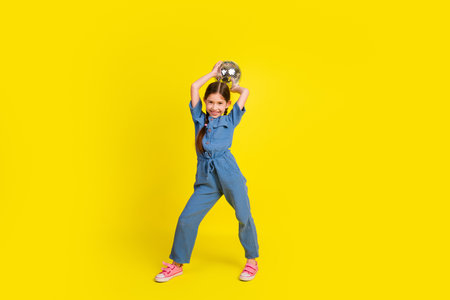 Smiling little girl holding a disco ball against a vivid yellow background, joyfully posing in casual attire.の写真素材