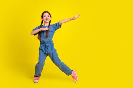 Happy young girl in a blue jumpsuit posing on a vibrant yellow background with joyful gestures and bright energyの写真素材