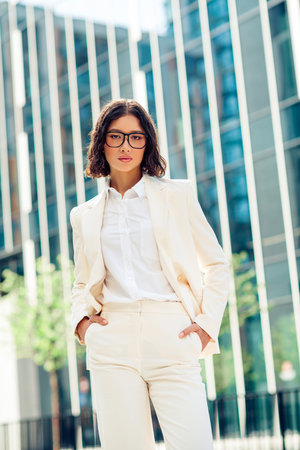 Elegant young businesswoman in professional attire standing confidently outdoors in a modern city environment during a sunny dayの写真素材