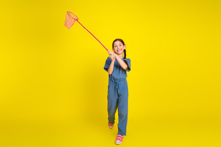 Cute young girl holding a butterfly net on a vibrant yellow background, wearing casual blue overalls and stylish braidsの写真素材
