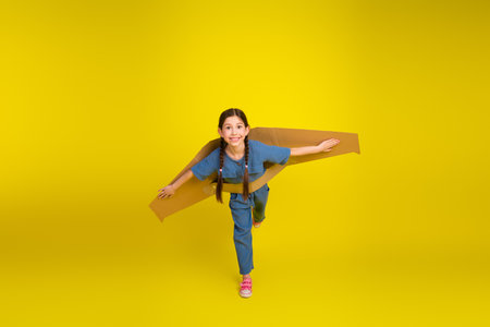 Happy young girl with handmade cardboard wings playing and smiling against a vibrant yellow backgroundの写真素材