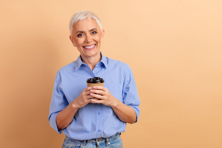Charming senior woman with short white hair holding a coffee cup, wearing a blue striped shirt, in a casual settingの写真素材