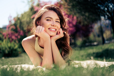 Lovely woman lying on grass in bright spring sunshine, enjoying leisure in a park, wearing yellow dress, smiling happilyの写真素材