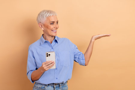 Mature woman in fashionable blue striped shirt holding smartphone and gesturing towards copy space against beige backgroundの写真素材