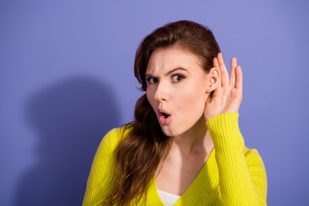 Young woman in bright yellow cardigan showcasing a listening expression against a vibrant purple backgroundの写真素材