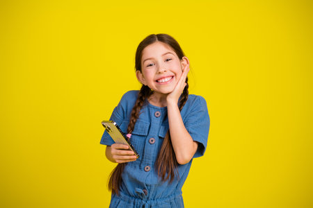 Adorable young girl with braids holding her phone on a vibrant yellow background exuding happiness...